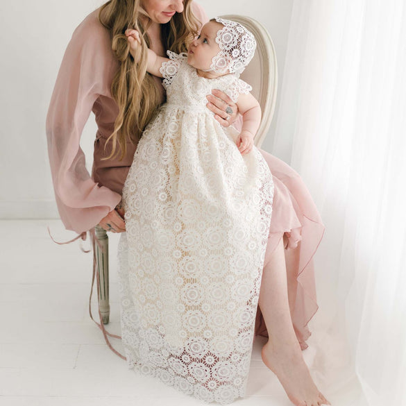 Baby wearing the Poppy Christening Gown and Bonnet, sitting on a woman's lap, showcasing the intricate floral lace overlay and long gown design in soft natural light.