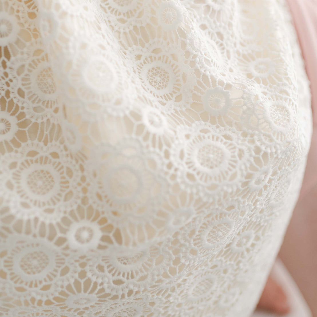 Close-up image of a Poppy Christening Gown & Bonnet with intricate floral and circular patterns, reminiscent of a Christening gown. The delicate material appears draped over a surface, showcasing the detailed and decorative design elements. Light and shadows create subtle variations in the texture.