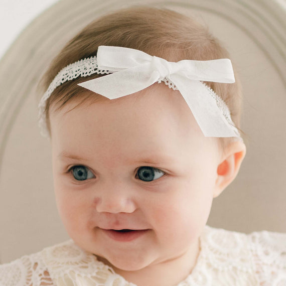 A baby with big blue eyes is wearing a white Poppy Headband with a bow. The child, dressed in a delicate Christening gown, is looking slightly to the side with a small smile. The background features a soft, neutral-toned surface.