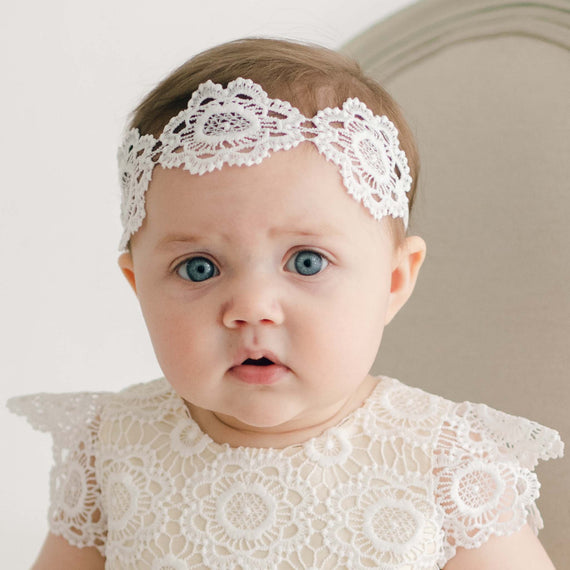 A baby with blue eyes and brown hair wears a white Poppy Lace Headband adorned with a light ivory flower lace, perfectly matching her delicate lace dress. Sitting against a light-colored background, the child gazes directly into the camera, with a neutral expression on her face. Handmade in the USA.
