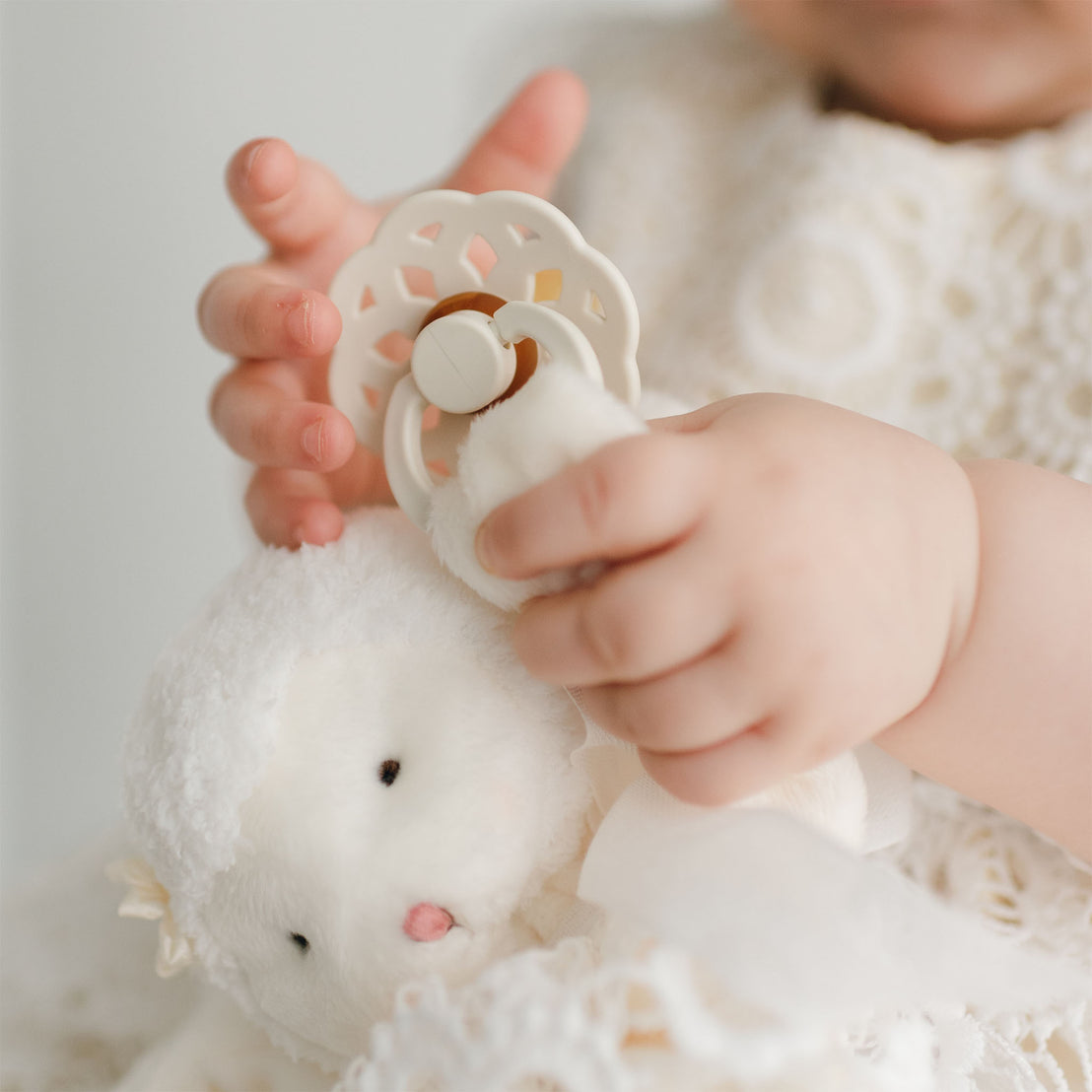 A baby dressed in white holds a Poppy Silly Lamb Buddy | Pacifier Holder while fiddling with a pacifier clip. The soft velour toy, also white, has a gentle face and a woolly texture, evoking a lamb or sheep. The baby's small hands are in focus, grasping the toy and pacifier delicately.