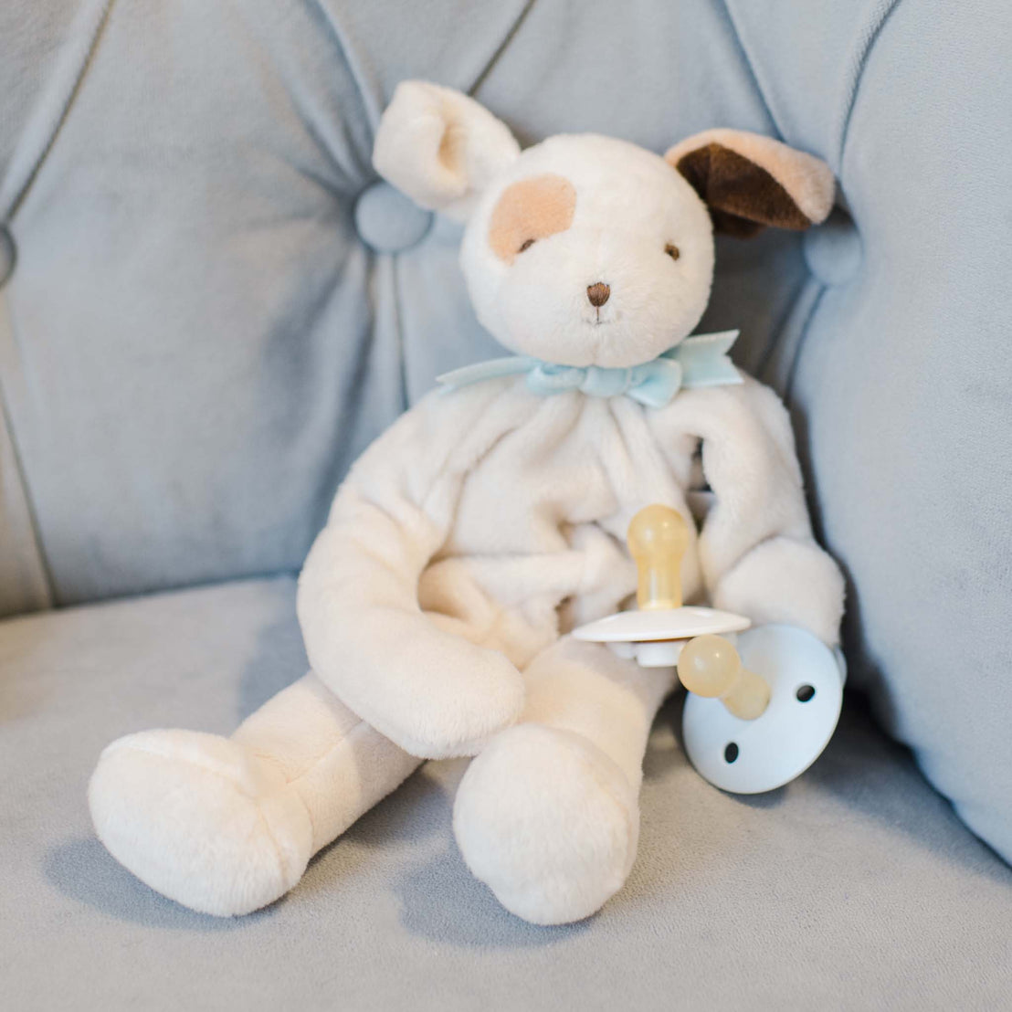 The Ezra Powder Blue Silly Puppy Buddy stuffed animal sitting on a couch. This stuffed animal serves as a pacifier holder