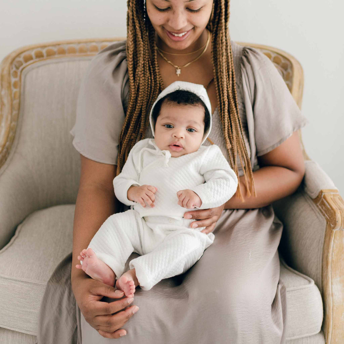 Baby girl sitting with her mother and wearing the Ivory Quinn Wrap Top & Pants. Crafted with extra soft quilted cotton and trimmed with a delicate ivory lace across the bodice and sleeves. She is also wearing the Ivory Quinn Quilted Cotton Bonnet.