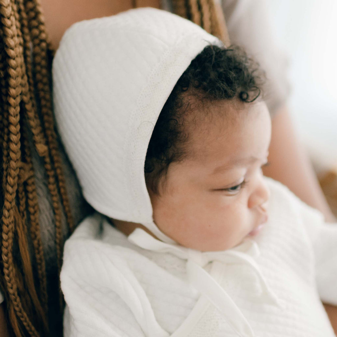 Baby girl with her mother and wearing the Ivory Quinn Quilted Cotton Bonnet. A traditional style bonnet that is lined in coordinating super soft pima cotton and trimmed in a light ivory cotton lace with soft pima cotton ties to secure the bonnet.