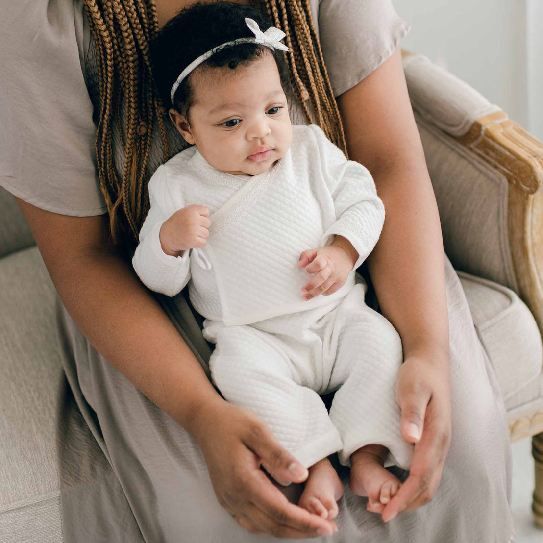 Baby girl sitting with her mother and wearing the Ivory Quinn Wrap Top & Pants. Crafted with extra soft quilted cotton and trimmed with a delicate ivory lace across the bodice and sleeves. She is also wearing the Quinn Lace Bow Headband.
