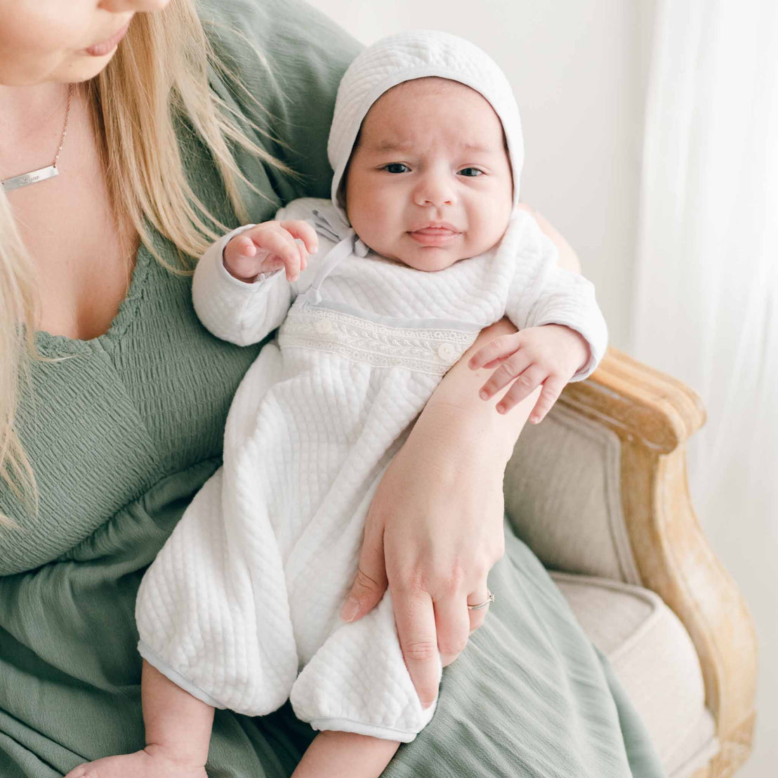 A newborn baby boy wearing a Harrison Quilted Bonnet made with soft quilted cotton in white and trimmed in a light blue linen, as well as the Harrison Newborn Romper 