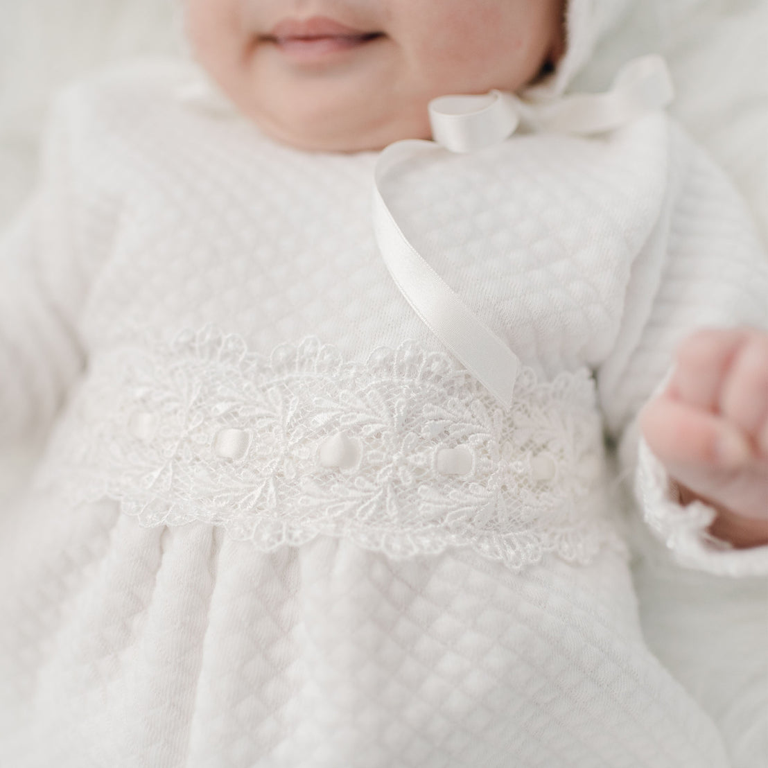 Close-up of a baby lying down, wearing a Madeline Quilted Newborn Romper with delicate lace and pearl detailing, perfect as an heirloom baby gift. 