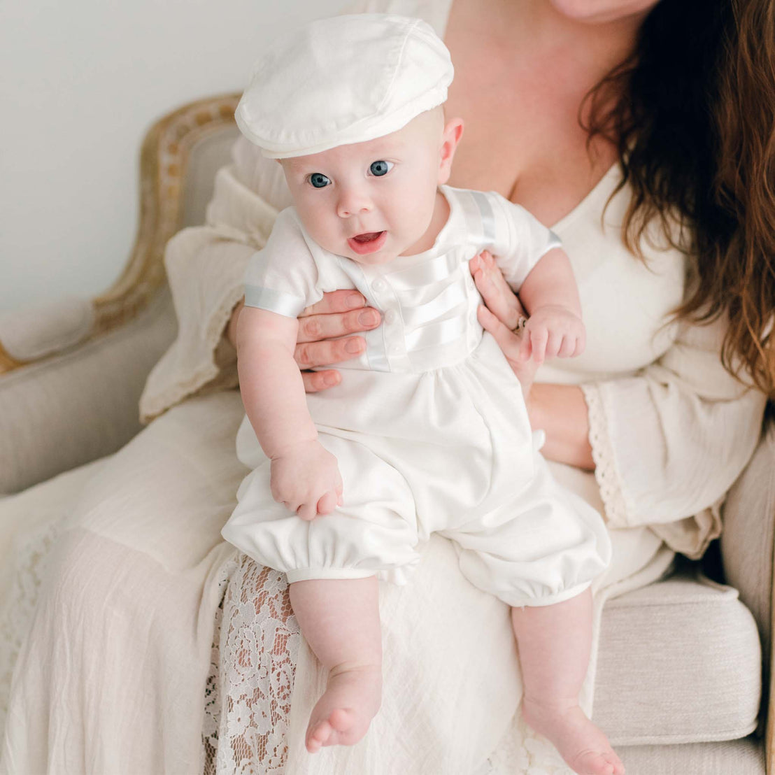 Baby boy sitting on his mother's lap and wearing the Owen Linen Romper featuring silk ribbon in ivory and light blue across the front bodice and sleeves