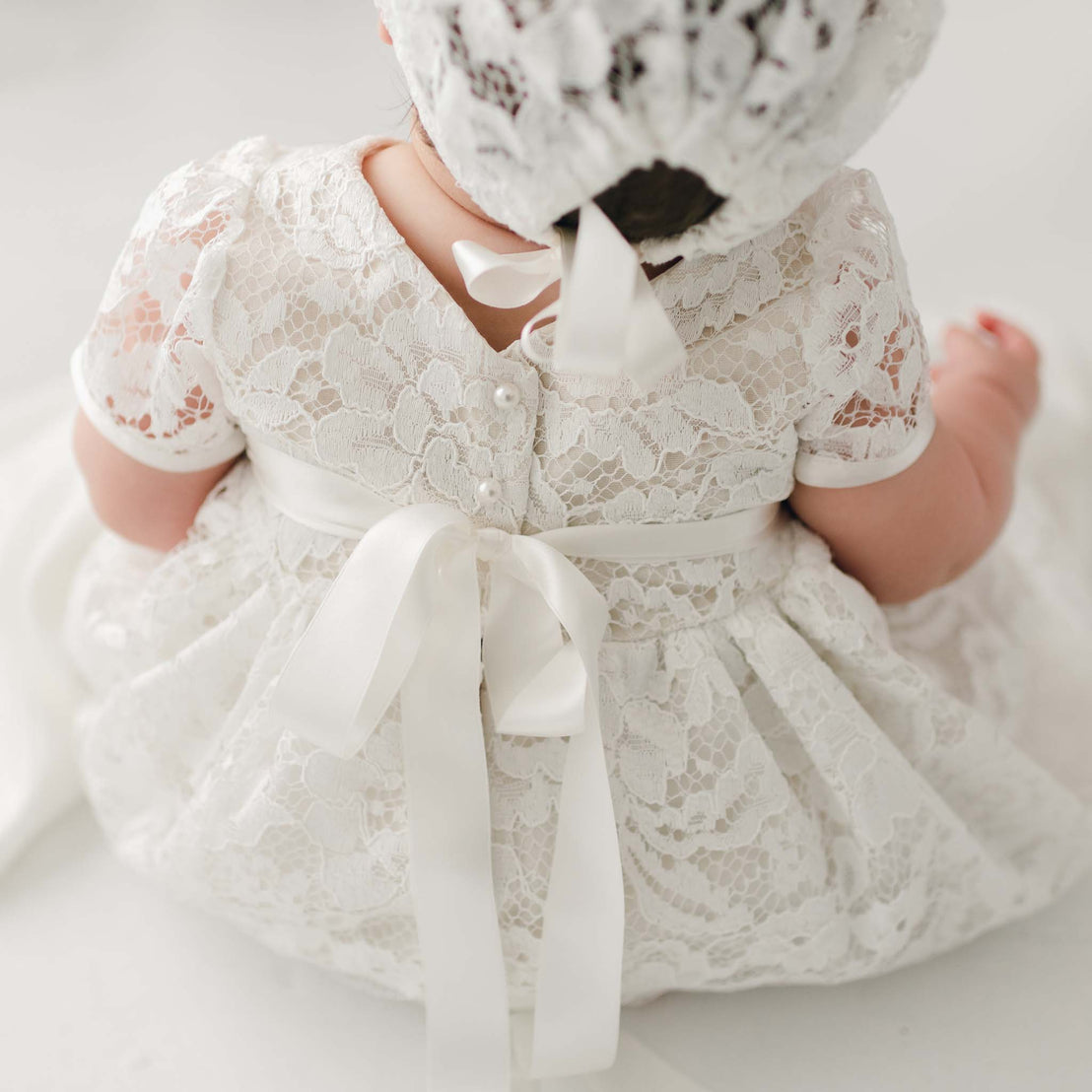 A baby wearing a delicate white Rose Christening Gown & Bonnet, viewed from behind, focusing on the details of the lace fabric and silk ribbon.