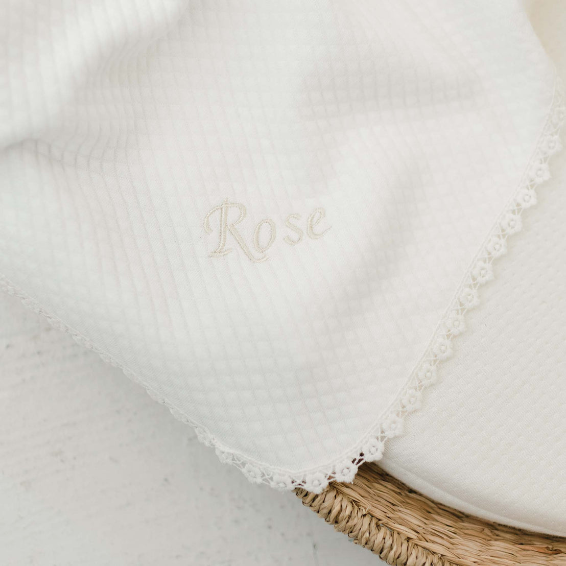 Close-up of a traditional white personalized blanket with the name "rose" embroidered in elegant script, framed by delicate lace trim, resting on an upscale wicker basket against a white background.