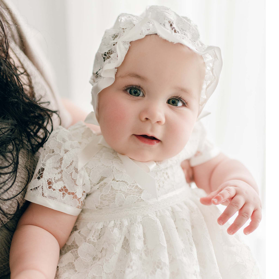 A close-up of a baby girl wearing a white lace Rose Christening Gown & Bonnet with pearl style buttons, looking directly at the camera with bright blue eyes and a subtle smile.