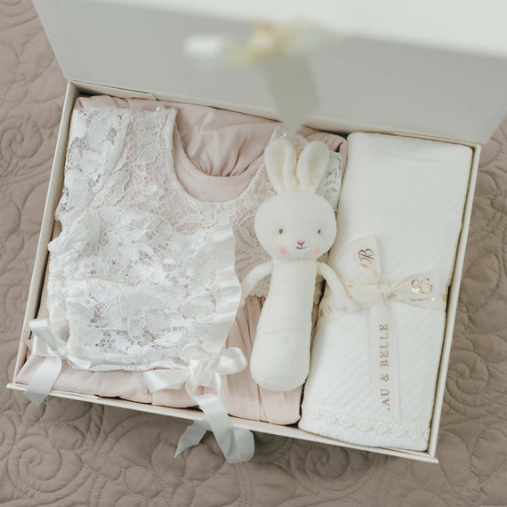 An open white gift box contains a blush pima cotton baby dress with a lace overlay, a matching lace bonnet, a white plush toy rabbit, and a cream-colored swaddling blanket with a ribbon and tag that reads "Rose Newborn Gift Set - Save 10%." The box is delicately placed on a quilted surface.