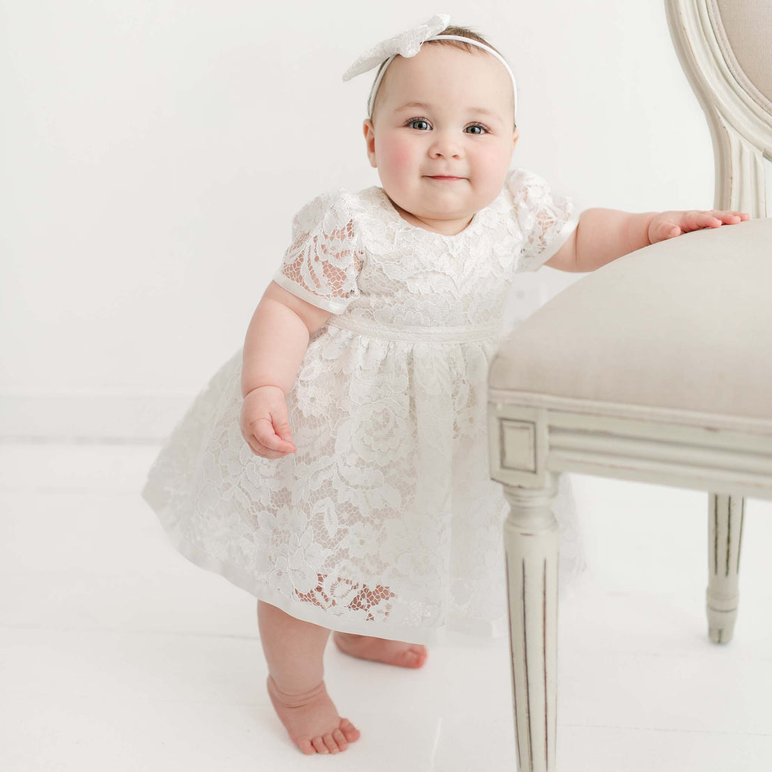 A baby girl in the Rose Romper Dress and Rose Bow Headband stands next to a beige chair in a bright room, smiling at the camera.