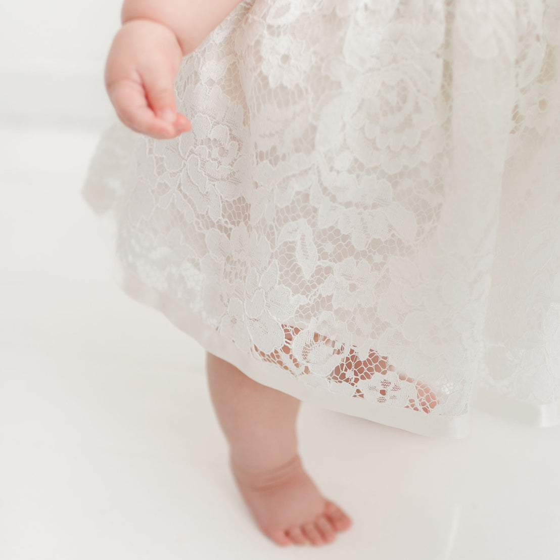 Close-up of a baby's lower body wearing a Rose Romper Dress, showing one bare foot and the hem of the dress. The background is plain white, emphasizing the texture of the lace.