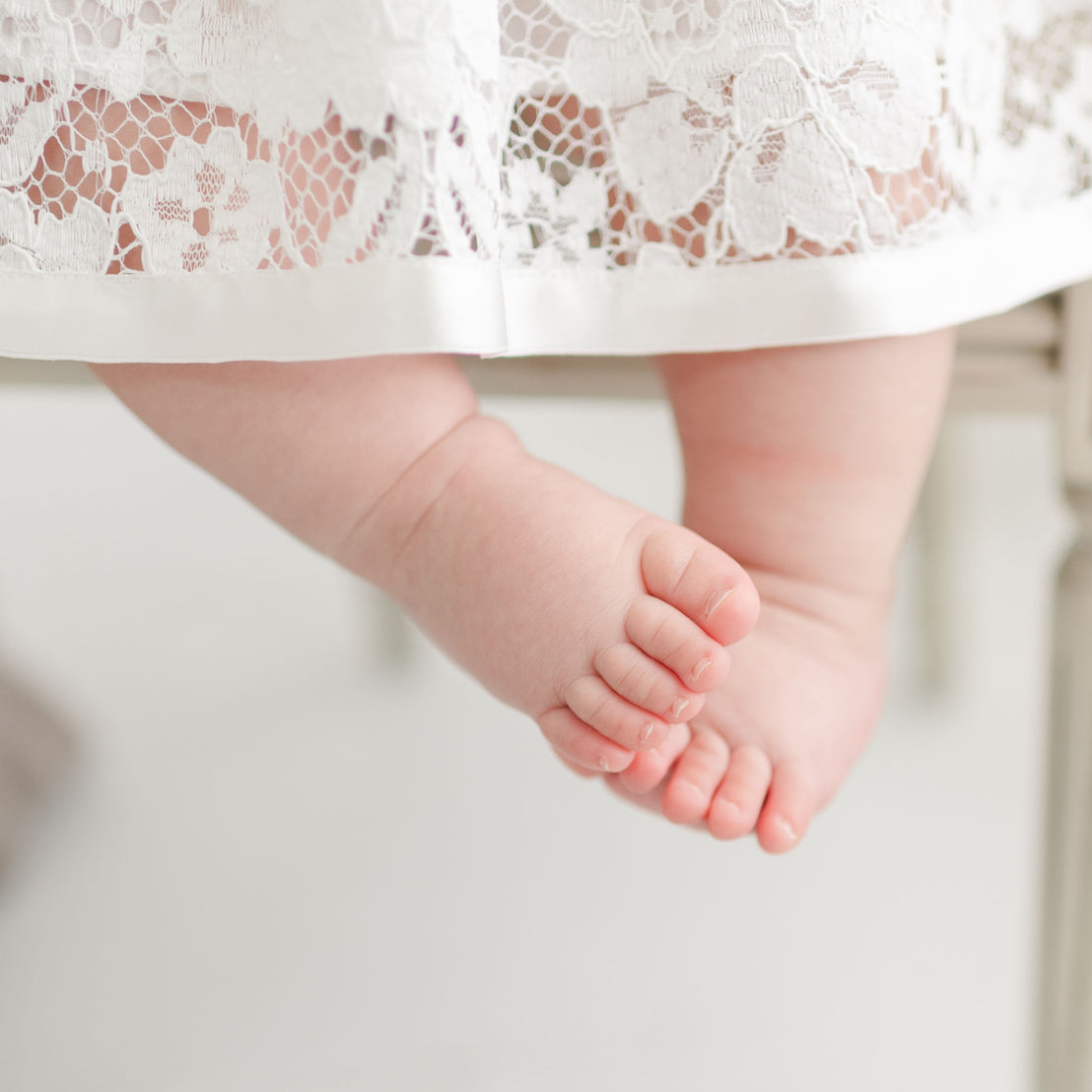 Close-up of a baby's bare feet dangling beneath a Rose Romper Dress skirt, with a blurred light background emphasizing the delicate texture and softness of the scene.