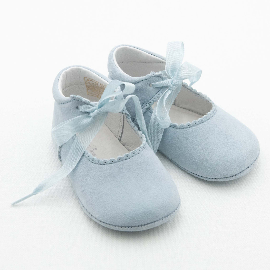 A pair of light blue baby shoes made of soft fabric, featuring delicate ribbon ties and scalloped edges around the opening. These Isla Suede Tie Mary Janes are positioned slightly overlapping on a white background.