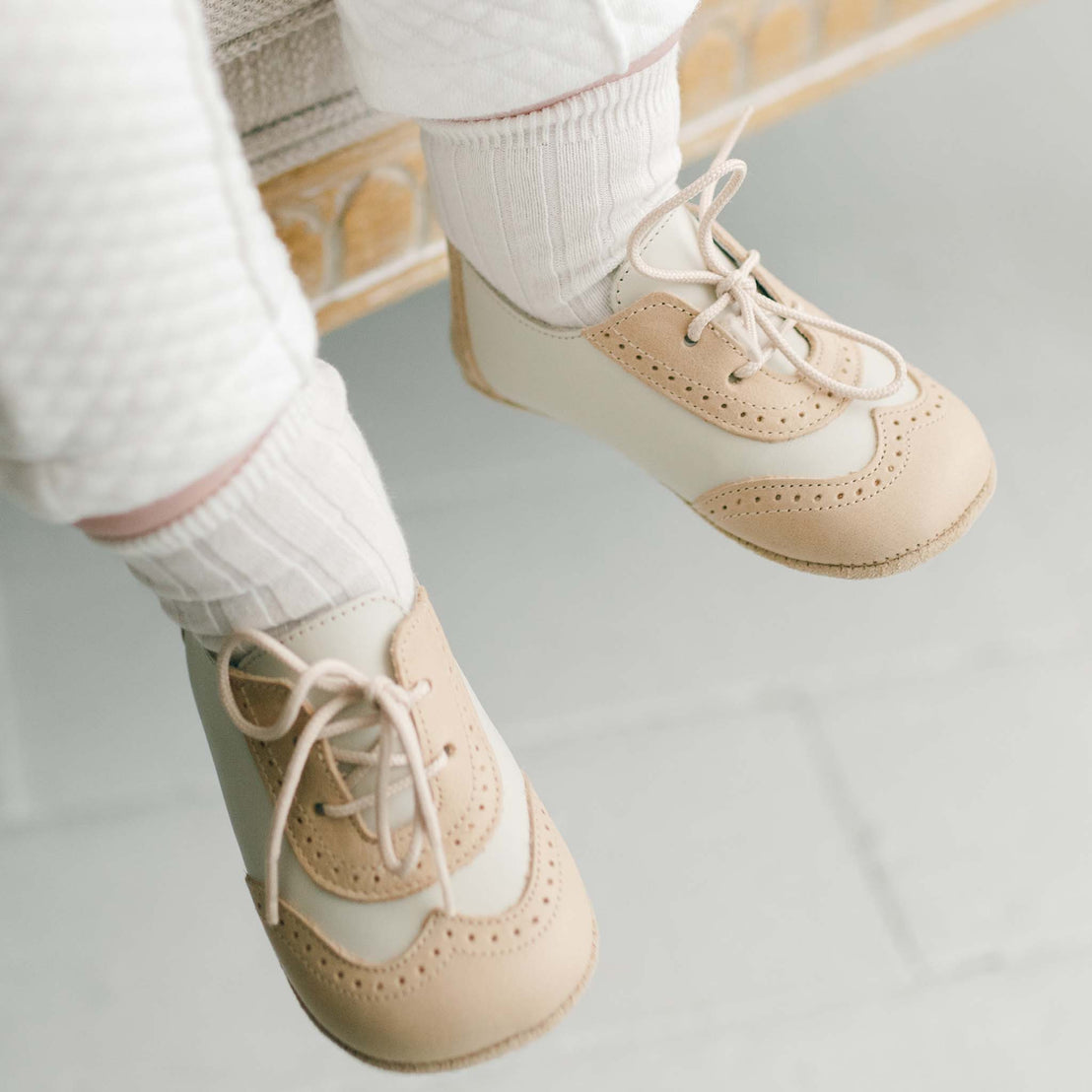Baby wearing a pair of the Beige and Ivory Wingtip Shoes made with a tan and ivory matte leather with tan suede soles and detailed edging