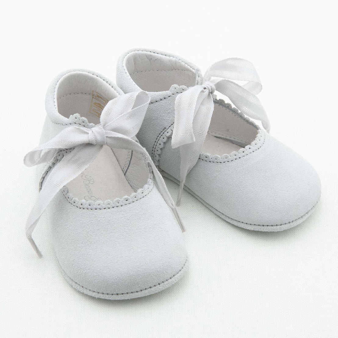 A pair of Girls Suede Tie Mary Janes with decorative white stitching and satin ribbon ties, displayed on a soft white background.