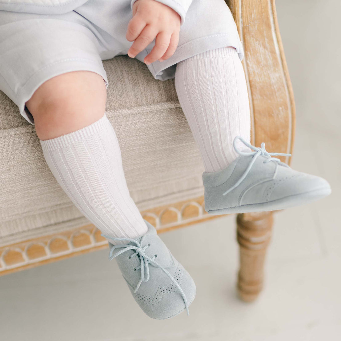Baby boy wearing the Harrison Suede Shoes crafted from a super soft, light blue suede with detailed edging. The boy is also wearing knee high white socks