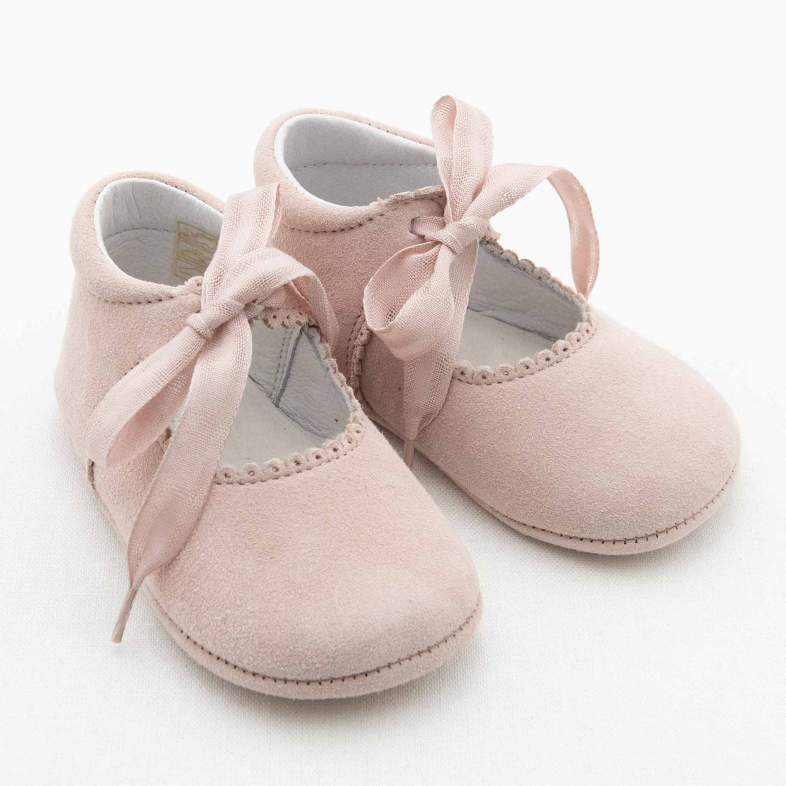 A pair of Emily Suede Tie Mary Janes, crafted in a delicate baby pink suede with scalloped edges and adorned with matching pink ribbon laces, sit side by side on a white background. These handmade shoes from Baby Beau & Belle have a soft, delicate appearance suitable for an infant.