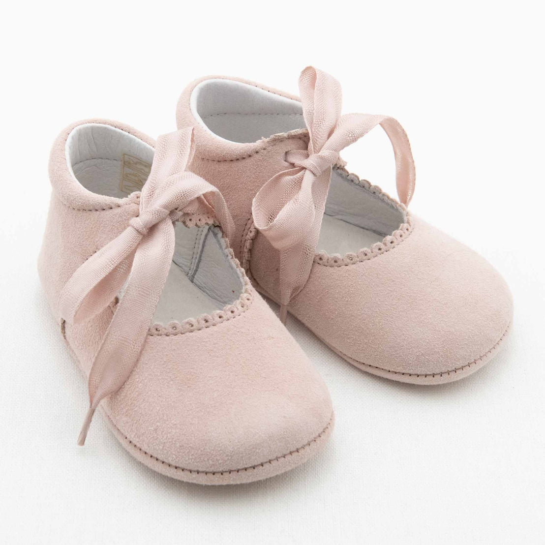 A pair of June Suede Tie Mary Janes from Baby Beau & Belle is seen. Crafted from blush suede, each shoe features a delicate satin bow tied at the front, scalloped edges around the opening, and a rounded toe design. The interior lining appears soft and comfortable, perfect for an infant.