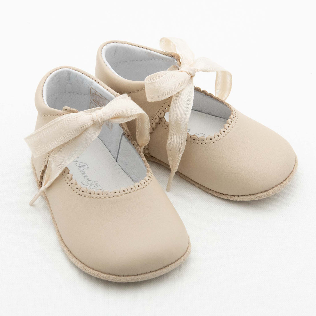 A pair of Tan Tie Mary Janes baby shoes with scalloped edges and a soft white interior. These Baby Beau & Belle shoes feature ivory satin ribbon ties and are displayed on a white surface.