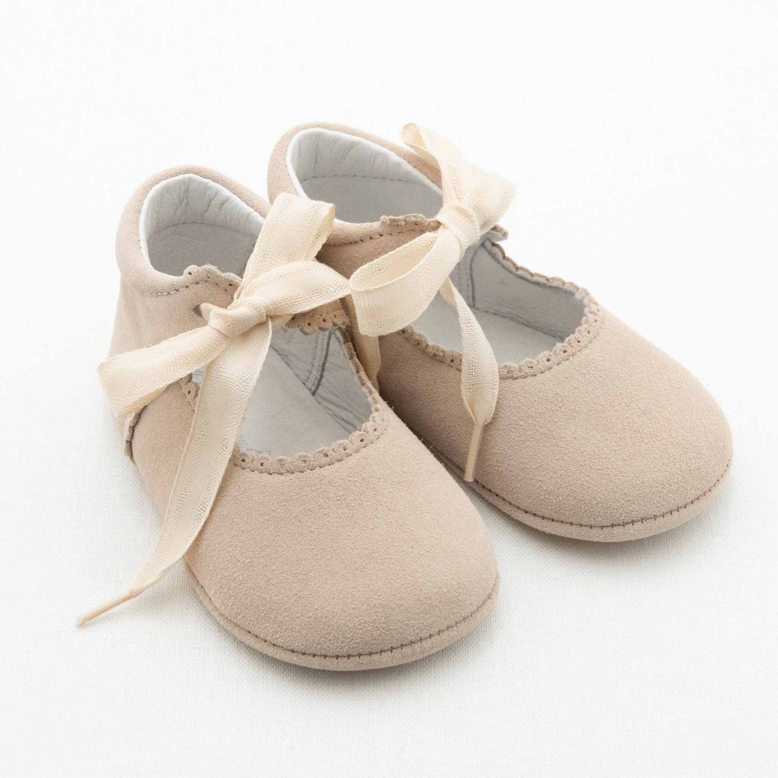 A pair of Kristina Tie Mary Janes from Baby Beau & Belle, light beige and soft, with scalloped edges and matching ribbon bows, are displayed on a white background. These handmade baby shoes, made of a suede-like material, exude an elegant appearance reminiscent of delicate Mary Janes.