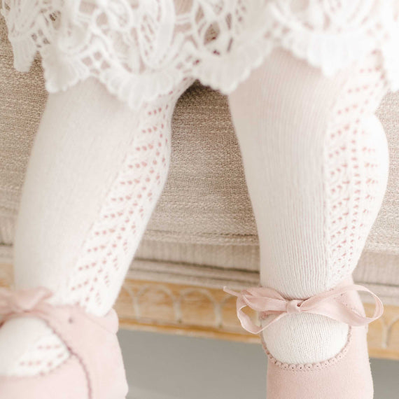 Close-up of a child's legs wearing white, lace-trimmed socks and Side Openwork Tights with ribbons tied around the ankles, feet dangling off the edge of an upscale, cushioned bench