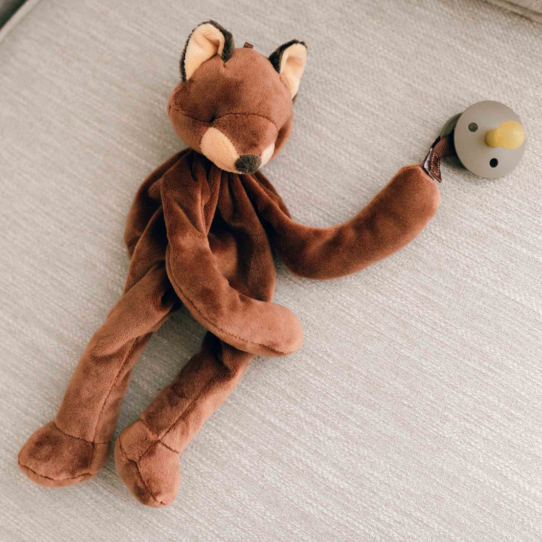 A Silly Fox Buddy pacifier holder with a rattle attached to its tail, lying on an upscale light grey fabric. The heirloom-quality toy is soft brown, and the rattle is green and white. Suitable
