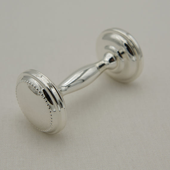 A silver rattle with circular faces and a decorative beaded border on one side, resting on a light beige textured fabric background.