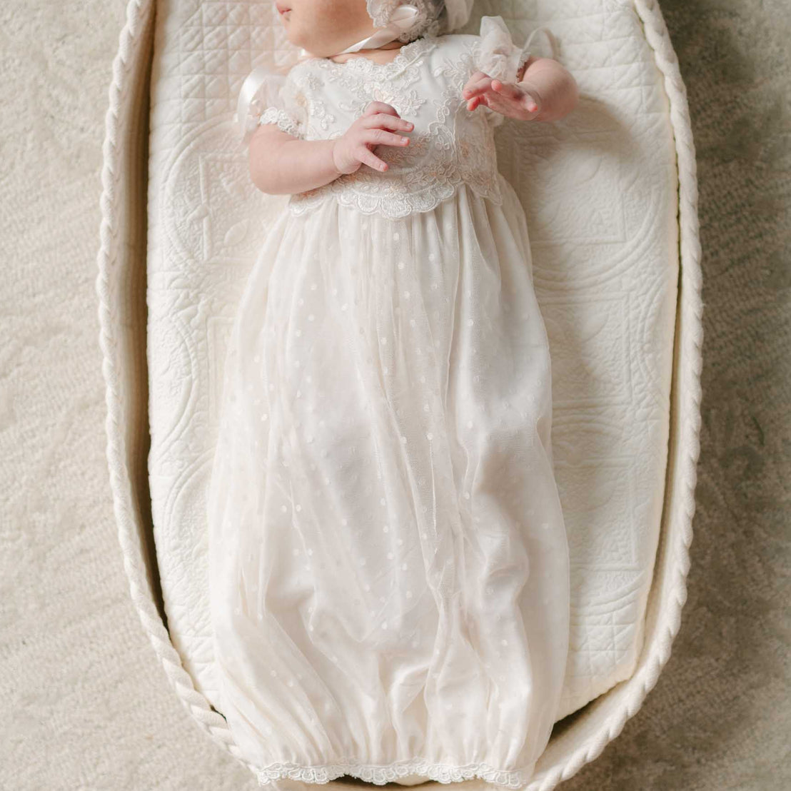 A newborn lays in a woven bassinet, dressed in the Sophia Layette with a matching lace bonnet, surrounded by soft textures.