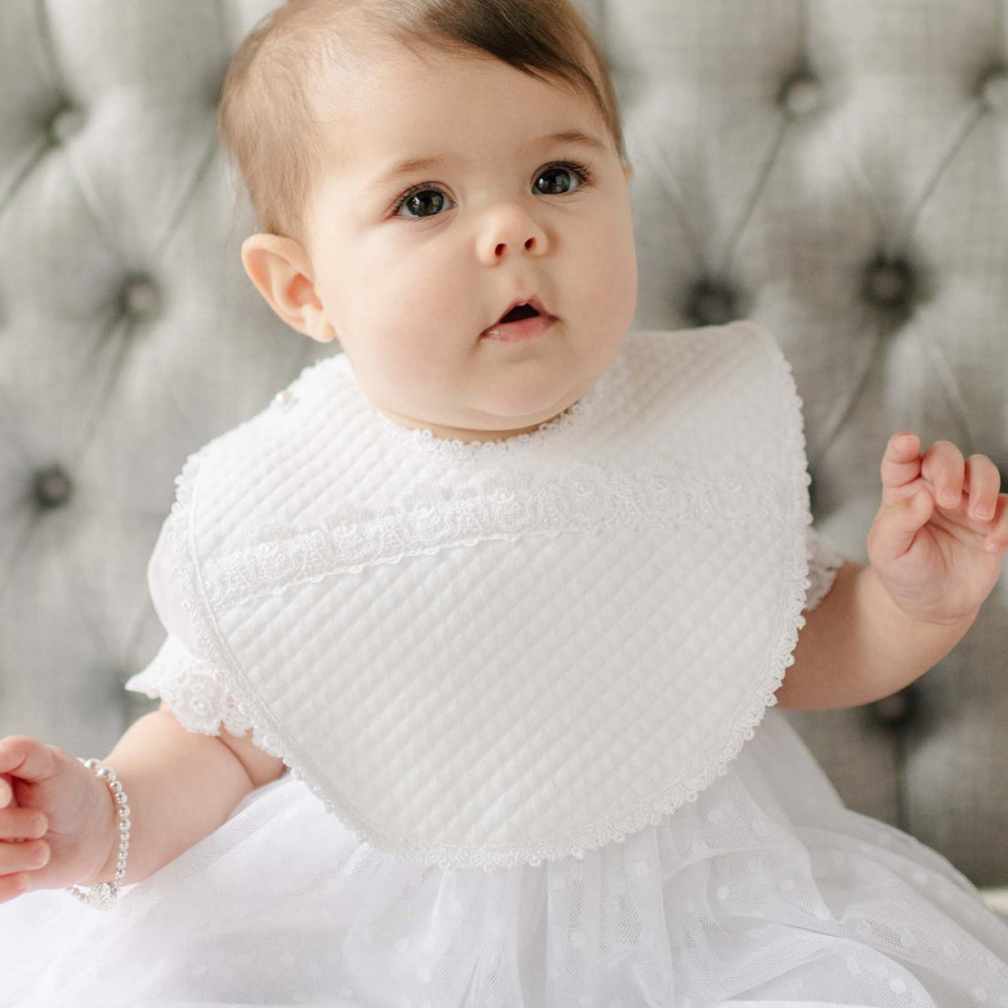 Baby wearing the Stella Bib against a gray tufted background. Details of the bib show a quilted cotton with a white edge lace trim. Perfect for unique baby gifts.