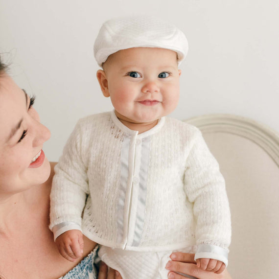 Baby boy smiling with his mother and wearing the Owen Knit Sweater made from a soft knit in ivory and trimmed with ivory linen and blue silk ribbon
