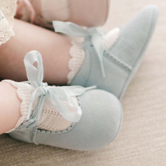 A baby wearing the blue Thea Suede Tie Mary Janes with ribbon ties and ruffled Pattern Socks, sitting on a beige textured surface.