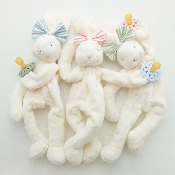 Three Thea Bunny Buddy toys, each wearing a miniature version of the Thea Bow Headband in blue, pink, green and holding a matching pacifier, lying on a white background.