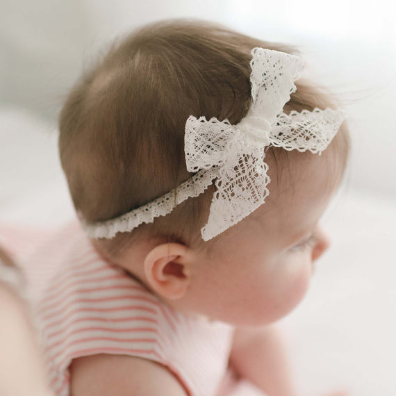 A profile view of a baby wearing the Thea Lace Headband, dressed in the pink Thea Wrap Dress, softly illuminated by natural light.