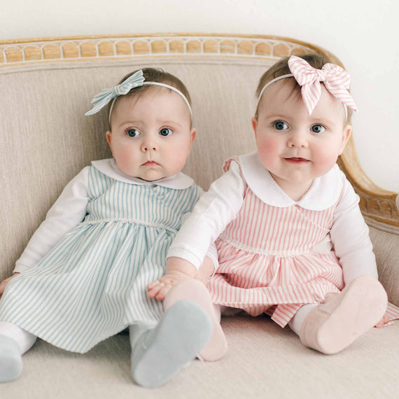 Two twin baby girls in the Thea Wrap Dresses, with white linen collar onesie underneath, and matching Thea Bow Headband sitting on a beige sofa, one looking curious and the other smiling at the camera.