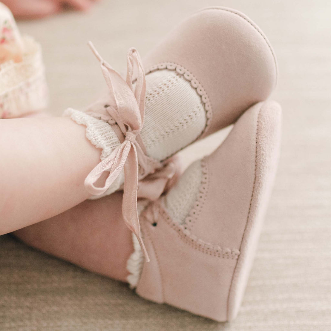 Close-up of a baby's feet wearing Thea Suede Tie Mary Janes with ribbon ties and white frilly socks, resting on a soft surface for their first birthday dress.