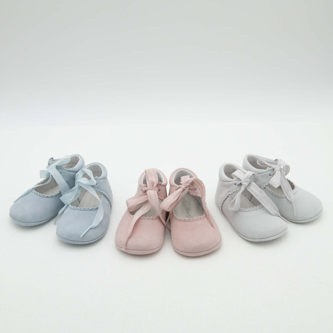 Three pairs of Thea Suede Tie Mary Janes in blue, pink, and gray, displayed in a row on a light background. Each pair features ribbon ties and decorative edges.