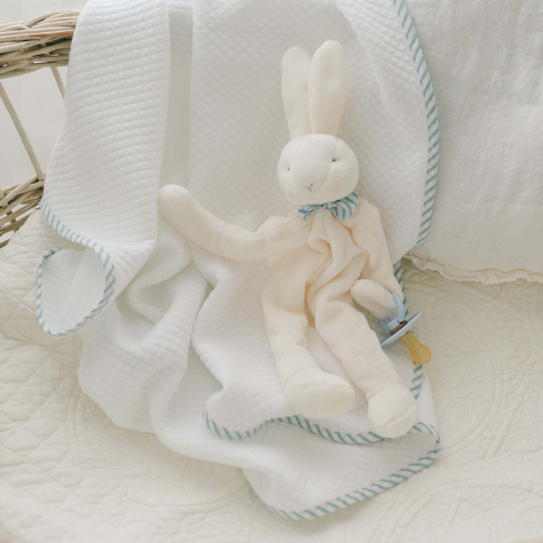 A plush white Theodore Bunny Buddy, crafted from soft velour with elongated limbs and a bow tie, is seated on a soft, white quilted blanket adorned with blue striped edges. The blanket is draped over a wicker basket, creating an ideal baby gift. Next to the bunny is a coordinating blue pacifier holder.