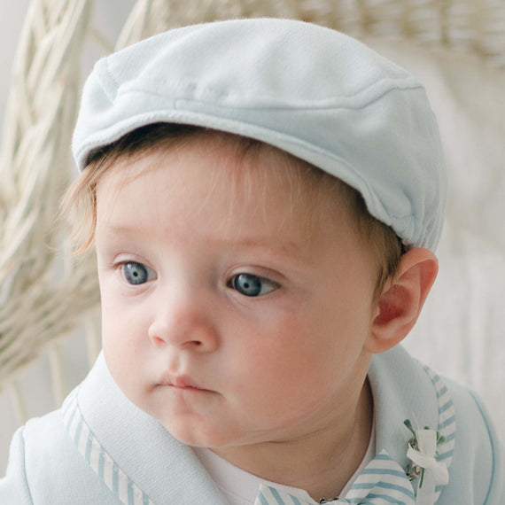 A baby with light brown hair and blue eyes is wearing a light blue Theodore Newsboy Cap and a matching christening outfit made of French Terry Cotton. The baby is looking slightly to the side, with a neutral expression. The background includes a white woven chair, creating a soft, pastel ambiance.