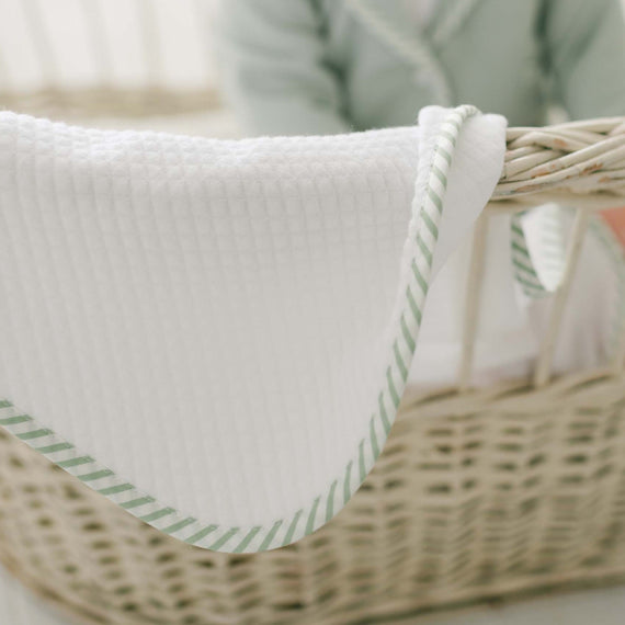 A baby wearing a green sweater is sitting in a wicker cradle. The focus is on the Theodore Personalized Blanket, crafted from 100% cotton, featuring a green striped trim draped over the side of the cradle. The baby's face is not visible.