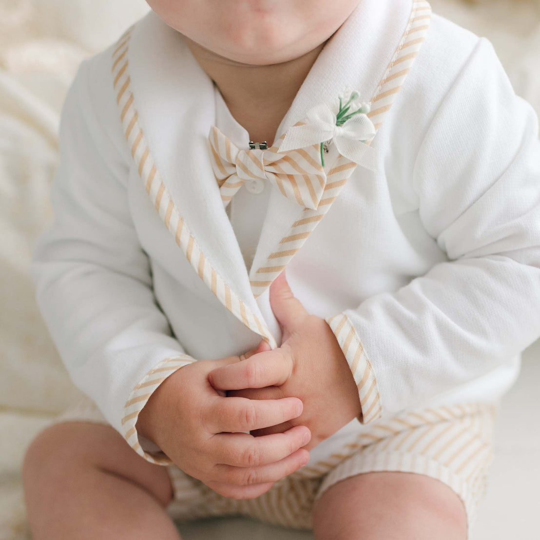 A baby is dressed in a white outfit with beige striped accents, including the Theodore Bow Tie & Boutonniere that features a handmade bow tie and trim. The baby’s hands are holding the lapel of the jacket, adorned with a boutonniere secured by a locking pin. Only the lower part of the baby’s face is visible.