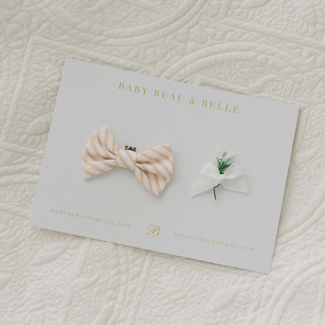 A card labeled "Theodore Bow Tie & Boutonniere" showcases a handmade bow tie crafted from beige and white striped cotton, accompanied by a small white flower with a ribbon. The card is placed on quilted white fabric, with "babybeauandbelle.com" and "christeninggowns.com" printed at the bottom.