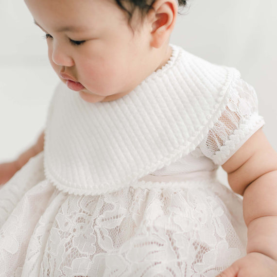 A close-up of a baby dressed in a delicate ivory floral lace Victoria Bib with a textured bodice, looking down thoughtfully.