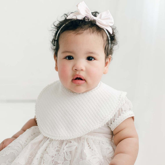 A baby girl with curly hair wearing a white Victoria Bib and a bow headband sits on a white background, looking directly at the camera.