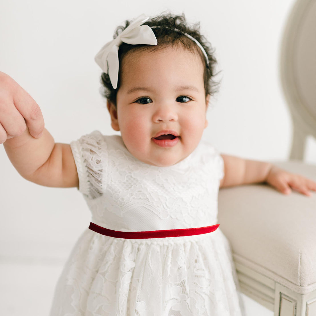 Baby in a white dress with a red belt and bow headband, sitting on a light-colored surface.