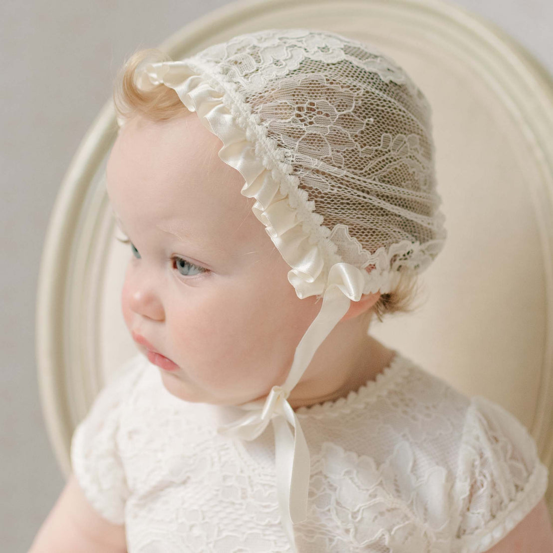 Close up photo of a baby wearing the Victoria Lace Bonnet against a neutral background.Details show the ivory floral lace and ivory silk ribbon that fits the face. Part of the Baby Beau & Belle Baptism Gown Girl.