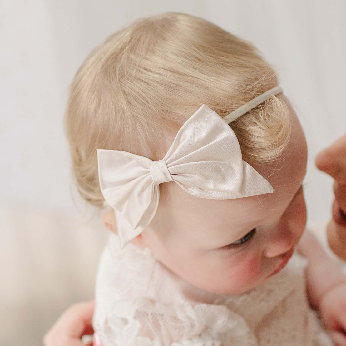 Baby wearing the Victoria Silk Bow Headband with a soft focus background. Detail shows a dupioni silk bow in pink champagne. Perfect for unique baby gifts.