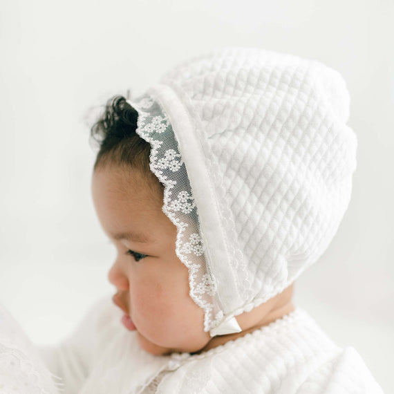 A close-up of a baby wearing a delicate Victoria Quilted Cotton Bonnet adorned with floral lace, focusing thoughtfully off to the side. The baby is dressed in a white outfit, providing a soft, pure appearance.